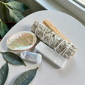 May include: An arrangement of spiritual items including a white sage bundle, a selenite wand, a clear quartz crystal, a palo santo stick, and an iridescent abalone shell. Green leaves are arranged around the items on a gray surface.