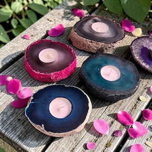 May include: A collection of four circular agate stone candle holders, each featuring a polished, colorful center and a natural, rough-textured outer edge. Each stone holds a small, round, white tea light candle in its center, displaying shades of pink, teal, and dark blue.