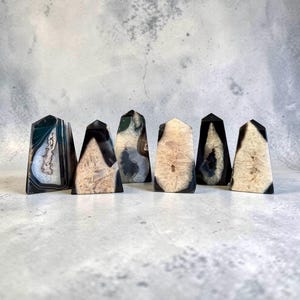 Puede incluir: Una colección de siete torres de cristal de ágata. Las torres presentan una variedad de colores, incluyendo negro, blanco, beige y verde. Cada torre tiene un patrón y forma únicos, con un acabado pulido. Los cristales se exhiben sobre una superficie gris claro.
