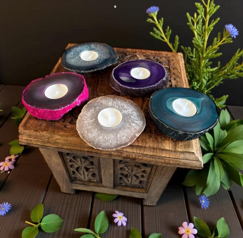 May include: Five circular tea light candle holders made from polished agate slices in shades of blue, purple, pink, and grey. Each holder features a central circular cutout containing a small white wax tea light candle, showcasing natural stone banding.
