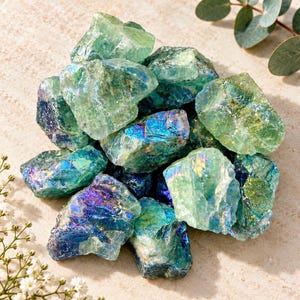 May include: A collection of raw, iridescent fluorite crystals featuring a vibrant spectrum of green, blue, and purple hues. These jagged, translucent mineral stones exhibit a reflective, metallic sheen across their multifaceted, uneven surfaces.