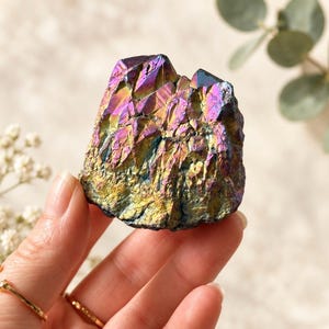 May include: A raw, iridescent crystal cluster featuring a vibrant spectrum of metallic colours including purple, blue, gold, and green. The mineral specimen has a jagged, multi-faceted surface with sharp, crystalline points and a dense, textured structure.