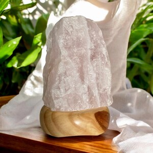 May include: A rose quartz crystal lamp in a pale pink hue, mounted on a natural wooden base. The crystal has a rugged, textured appearance. The lamp is displayed on a wooden surface, with a white cloth and green foliage in the background.