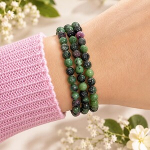 May include: A triple-strand beaded bracelet featuring polished round stones in shades of forest green, dark charcoal, and vibrant magenta. The beads are strung on an elastic cord, creating a layered look with varied natural stone textures and colors.