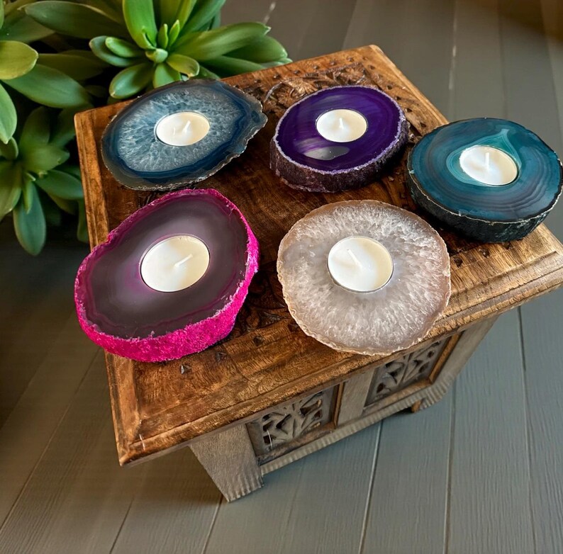 May include: Five circular agate stone candle holders with natural crystalline centers and polished edges. Each holder features a central circular cutout containing a small white tea light candle. The stones are colored in blue, purple, teal, magenta, and beige.