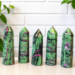 May include: Five polished crystal towers arranged in a row, each featuring a vibrant green base with distinct black speckles and streaks of deep magenta-pink throughout the stone, standing upright with a pointed hexagonal top and flat base.