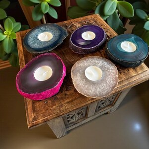 May include: Five circular agate stone candle holders with polished surfaces and natural rough edges. Each piece features a central circular cutout holding a small white tea light candle. The stones are colored in blue, purple, teal, pink, and beige.