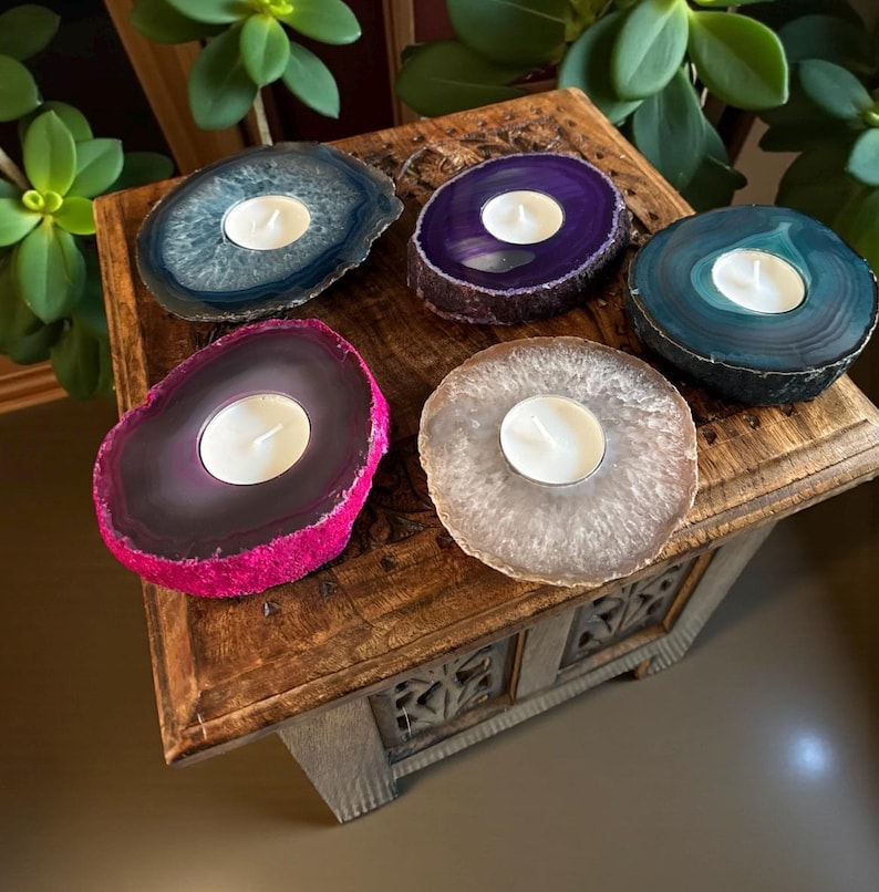 May include: Five circular agate stone candle holders with polished surfaces and natural rough edges. Each piece features a central circular cutout holding a small white tea light candle. The stones are colored in blue, purple, teal, pink, and beige.