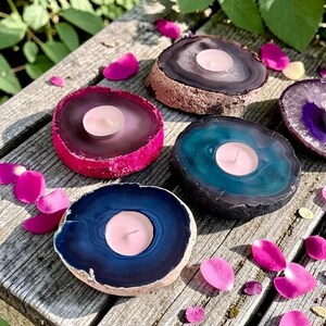 May include: Four circular agate stone candle holders in shades of dark blue, teal, magenta, and brown, each featuring a central circular cutout holding a small white tea light candle, arranged on a weathered wooden surface with scattered pink petals.