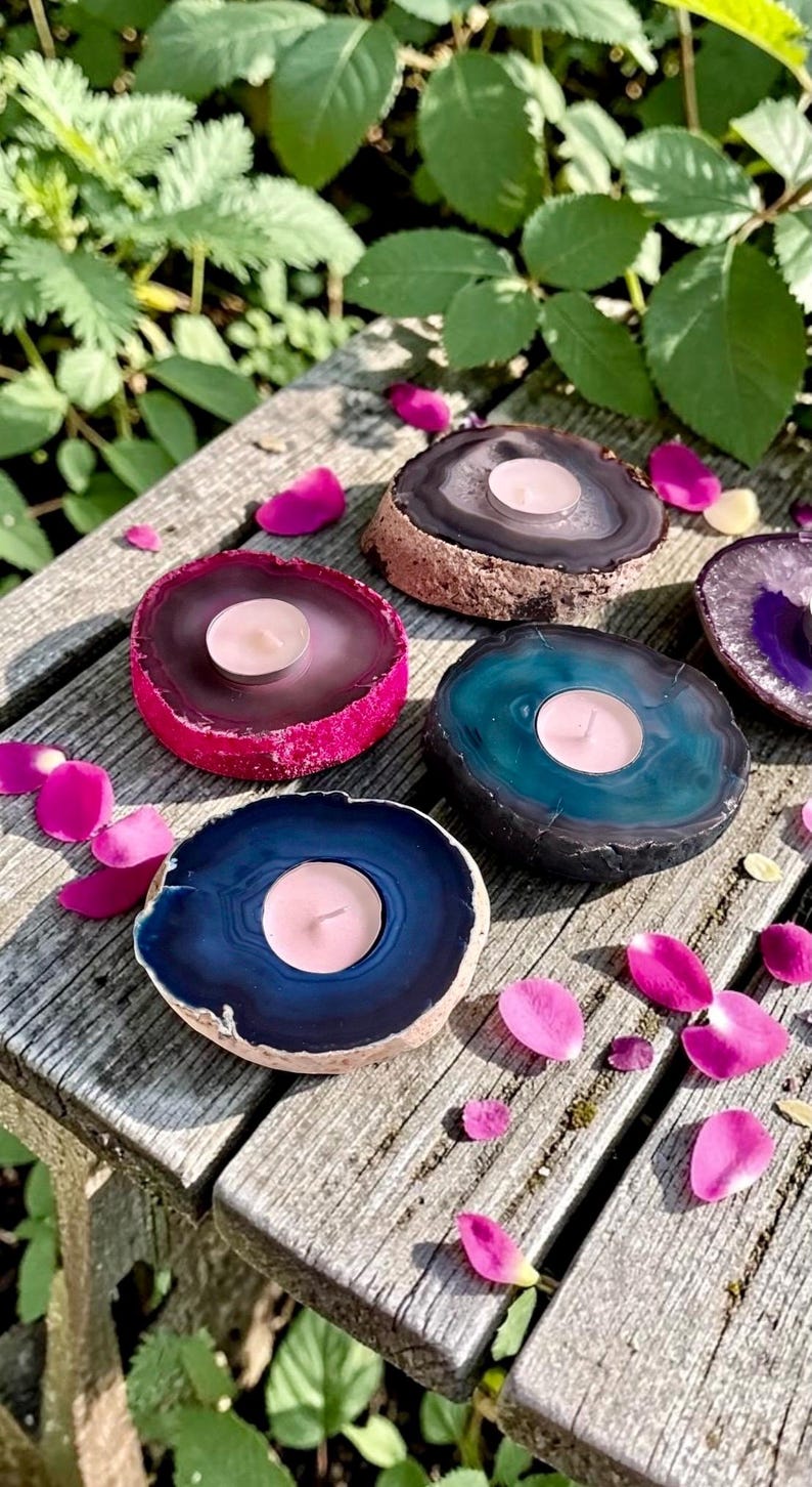 May include: Four circular agate stone candle holders in shades of dark blue, teal, magenta, and brown, each featuring a central circular cutout holding a small white tea light candle, arranged on a weathered wooden surface with scattered pink petals.