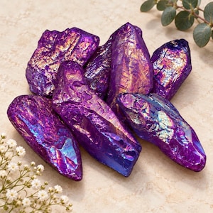 May include: A collection of six raw, jagged crystal points featuring a vibrant, metallic purple aura coating. The surfaces of these stones exhibit iridescent flashes of gold, blue, and magenta, creating a reflective, multi-faceted appearance.