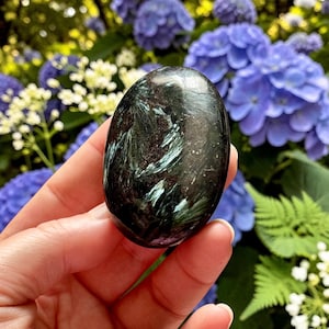 May include: A polished, oval-shaped seraphinite gemstone featuring a deep dark green base color with intricate, feathery, light green and silver chatoyant inclusions that create a shimmering, needle-like pattern across the smooth, reflective surface.