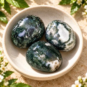 Puede incluir: Tres piedras de ágata musgosa pulidas con inclusiones de color verde oscuro y blanco translúcido que descansan dentro de un pequeño cuenco de cerámica de color blanco roto. Las piedras tienen una superficie lisa y redondeada.