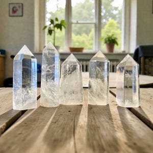 Może przedstawiać: Pięć przezroczystych kryształowych punktów kwarcu o różnych rozmiarach ułożonych na drewnianej powierzchni. Kryształy są przezroczyste z wewnętrznymi wtrąceniami, a każdy ma spiczasty wierzchołek. Kryształy są wystawione przed oknem z naturalnym światłem.