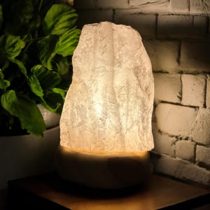 May include: A natural salt lamp with a textured, uneven surface, emitting a warm glow. The lamp sits on a light-coloured base. The lamp is placed on a wooden surface, with a brick wall and a green plant in the background.