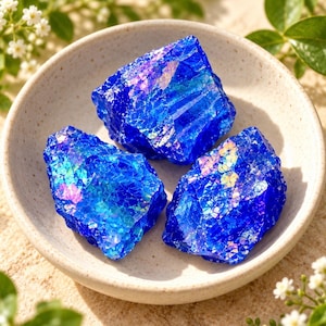 May include: Three rough, jagged pieces of vibrant blue glass with a crackled, iridescent finish are arranged inside a shallow, off-white ceramic bowl. The glass fragments feature sharp, angular edges and reflective surfaces.