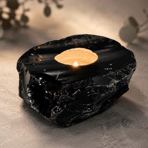 May include: A black, glossy, rock-like candle holder with a rough, uneven exterior texture and a smooth, circular indentation at the top. A small, lit tea light candle sits inside the centre, emitting a warm, soft glow from the hollow space.