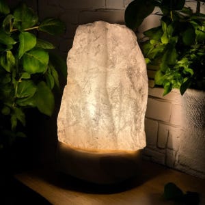 May include: A lit rock salt lamp with a textured, irregular surface. The lamp emits a warm, soft glow. The lamp sits on a rounded base, with green plants in the background. The lamp is a decorative item.