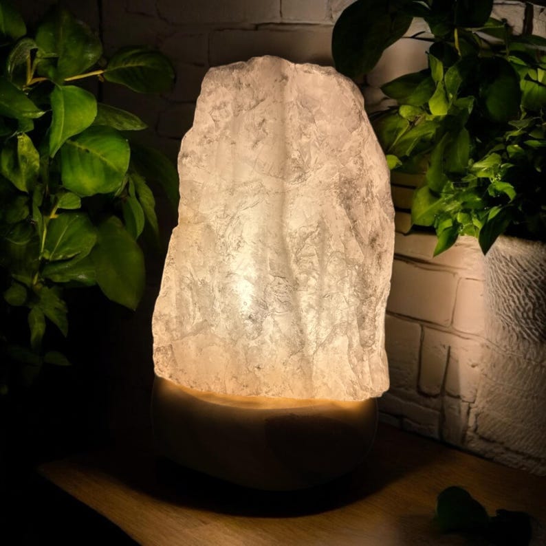 May include: A lit rock salt lamp with a textured, irregular surface. The lamp emits a warm, soft glow. The lamp sits on a rounded base, with green plants in the background. The lamp is a decorative item.