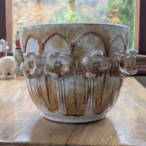 May include: A cream and brown ceramic planter. The planter has embossed floral designs around the rim and a row of flowers around the middle. Vertical lines decorate the lower portion of the planter. The planter is likely for indoor use.