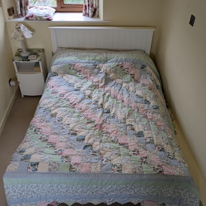 May include: A bed with a white headboard covered in a patchwork quilt featuring a variety of floral and gingham patterns in soft shades of pink, blue, green, and yellow. The quilt has a scalloped edge and covers the mattress and pillows.