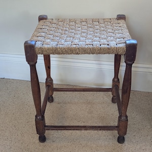 May include: A small, square wooden stool with a woven seat. The stool has four turned legs and a dark brown finish. The seat is made of woven natural fibres. The stool is a vintage or antique piece.