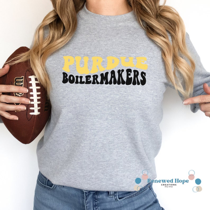 Caucasian female with football in hand wearing a stylish Purdue Boilermaker gray crewneck sweatshirt showcasing the SVG design available for instant download.