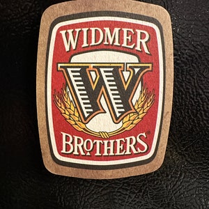 Puede incluir: Un posavasos de cerveza marrón y beige con el texto "Widmer Brothers" en letras negras y un logotipo "W" estilizado en negro con detalles dorados. El fondo es rojo.