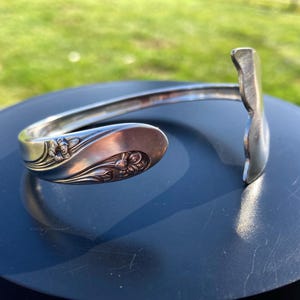 May include: A silver-toned metal cuff bracelet fashioned from a repurposed spoon. The bracelet features an open design with a polished finish and intricate embossed floral detailing on the curved ends, showcasing the original handle shape.