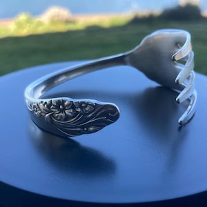 May include: A silver-colored bracelet crafted from a repurposed fork. The bracelet features a floral design on one end and the tines of the fork on the other. The bracelet is displayed on a dark surface.
