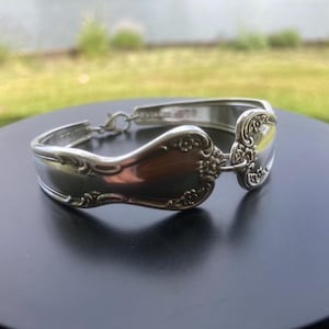 May include: Silver spoon bracelet with ornate floral detailing. The bracelet is made from repurposed silverware, featuring a decorative design on the spoon bowls and handles. The bracelet is connected with a small chain and clasp.