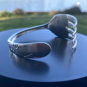 May include: A silver-colored bracelet crafted from a repurposed fork. The bracelet features a floral design on the handle and the tines of the fork form the open end. The bracelet is displayed on a dark surface.