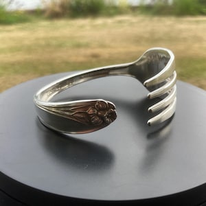 May include: A silver-toned metal cuff bracelet fashioned from a repurposed fork. The bracelet features a curved handle with an embossed floral motif at one end and the four tines of the fork bent outward at the opposite end to form the opening.