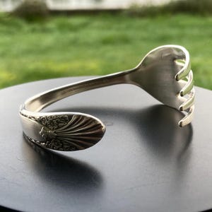 May include: A silver-colored bracelet crafted from a repurposed fork. The bracelet features the fork's tines at one end and the decorated handle at the other. The handle has a floral design. The bracelet is displayed on a dark surface.