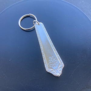 May include: Silver-colored metal keychain featuring a decorative spoon-shaped pendant. The pendant has an ornate floral design at the bottom and a textured border. The keychain ring is gold-colored.