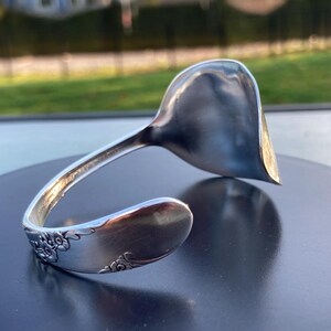 May include: A silver-toned metal cuff bracelet fashioned from a repurposed spoon. The bracelet features a curved, open-ended design with a smooth, polished bowl section at one end and a handle section decorated with embossed floral patterns at the other.