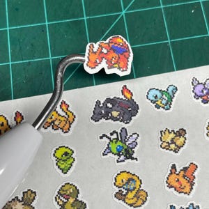 May include: A collection of pixel art stickers featuring various colorful creatures. One sticker is held by a metal tool. The stickers are on a white background, with a green cutting mat in the background.