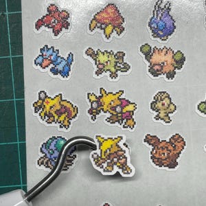 May include: A collection of colorful pixel art stickers featuring various creatures. The stickers are in a variety of colors, including yellow, blue, red, and green. A metal tool is lifting one of the stickers.