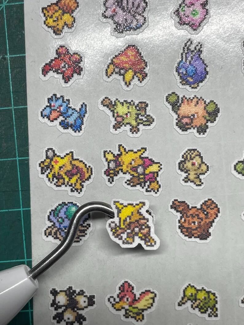 May include: A collection of colorful pixel art stickers featuring various creatures. The stickers are in a variety of colors, including yellow, blue, red, and green. A metal tool is lifting one of the stickers.