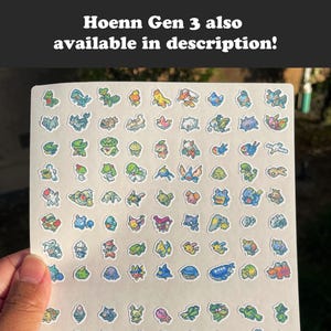 May include: A sheet of colorful stickers featuring various pixelated creatures. The stickers are arranged on a white background and held by a hand. The text "Hoenn Gen 3 also available in description!" is at the top.