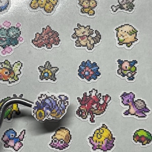 May include: A collection of pixelated stickers featuring various colorful creatures. The stickers have a white border and are arranged on a light gray surface. One sticker is held by tweezers, showcasing the detailed design.