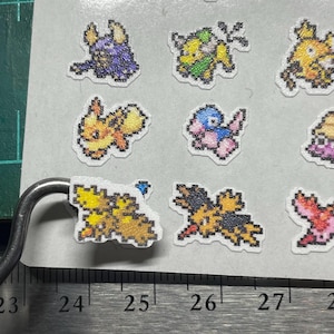 May include: Sheet of pixel art stickers featuring various colorful cartoon creatures. The stickers are on a white background and have a pixelated design. A metal ruler is in the foreground, with measurements in inches.