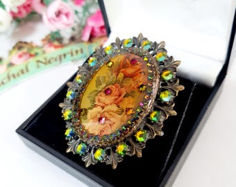 Può includere: Spilla ovale con un design floreale con rose nei toni del pesca e del giallo. La spilla è incorniciata da una filigrana color bronzo e circondata da cristalli iridescenti verdi e gialli. Riposa in un portagioie nero.