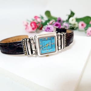 May include: A wristwatch with a square face featuring a bright turquoise dial, gold-tone hands, and numerals. The watch has a silver-tone metal frame with decorative beaded accents and a dark brown embossed leather strap with a crocodile texture.