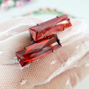 Puede incluir: Dos piezas decorativas rectangulares facetadas de vidrio o resina, engastadas en garras de metal color latón. Una pieza tiene un aspecto translúcido similar al ámbar con motas doradas, mientras que la otra es de un rojo rubí claro y profundo.