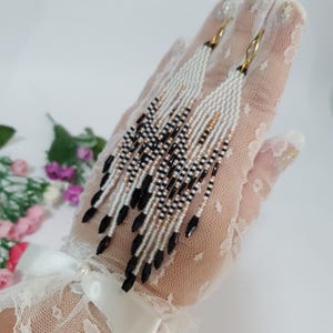 Puede incluir: Pendientes largos con cuentas con un patrón blanco, negro y dorado. Los pendientes tienen un diseño de flecos con cuentas negras en forma de lágrima en la parte inferior. Los pendientes se muestran sobre una superficie cubierta de encaje.