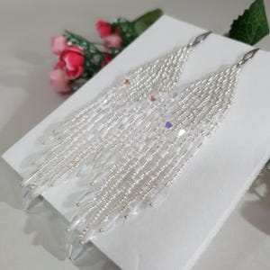 May include: A pair of long, dangling fringe earrings crafted from small, iridescent white and silver beads. The design features multiple cascading strands of varying lengths, finished with clear, faceted crystal-like beads at the bottom tips.