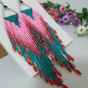 May include: A pair of long, beaded earrings with a triangular top and dangling fringe. The earrings feature a color gradient of pink, teal, and red beads, with iridescent red crystal accents. The earrings have silver-colored metal findings and a small green gem.