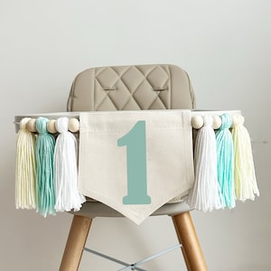 Op de afbeelding: Een hoge stoel met een witte stoffen banner waarop het getal "1" in teal groen staat. De banner is versierd met een rij houten kralen en kleurrijke garenkwastjes in geel, wit en teal groen.