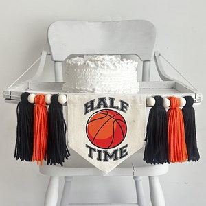 Może przedstawiać: Białe krzesełko do karmienia z białym ciastem ozdobionym falbankami i banerem z napisem "HALF TIME" i grafiką koszykówki. Baner jest ozdobiony czarnymi i pomarańczowymi frędzlami.
